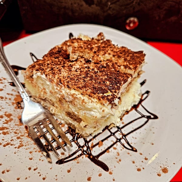 A slice of tiramisu with chocolate drizzle and cocoa powder on a white plate with a fork.
